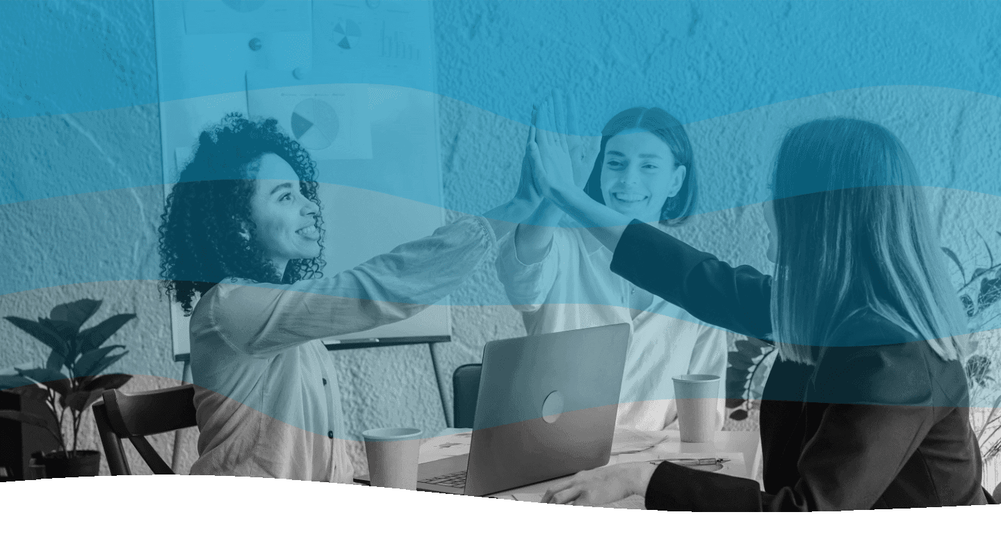Four people sitting at a wooden table in a bright, modern office, celebrating with a high-five; laptops, papers, and a small plant adorn the workspace.