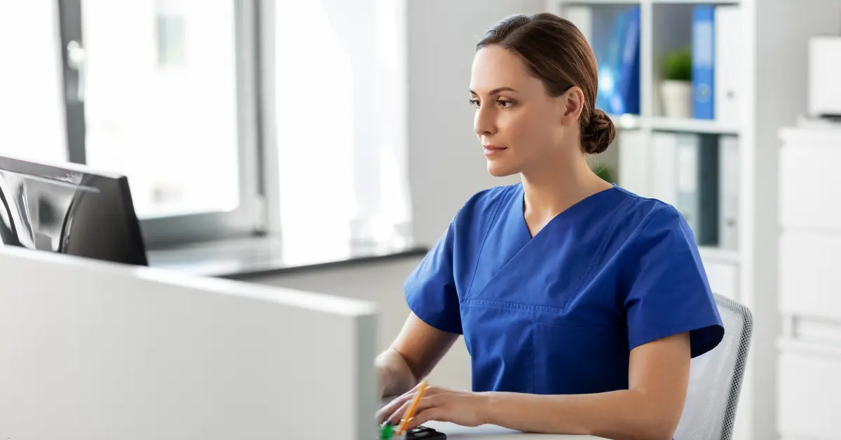 Nurse in blue scrubs working at a computer in a bright medical office.