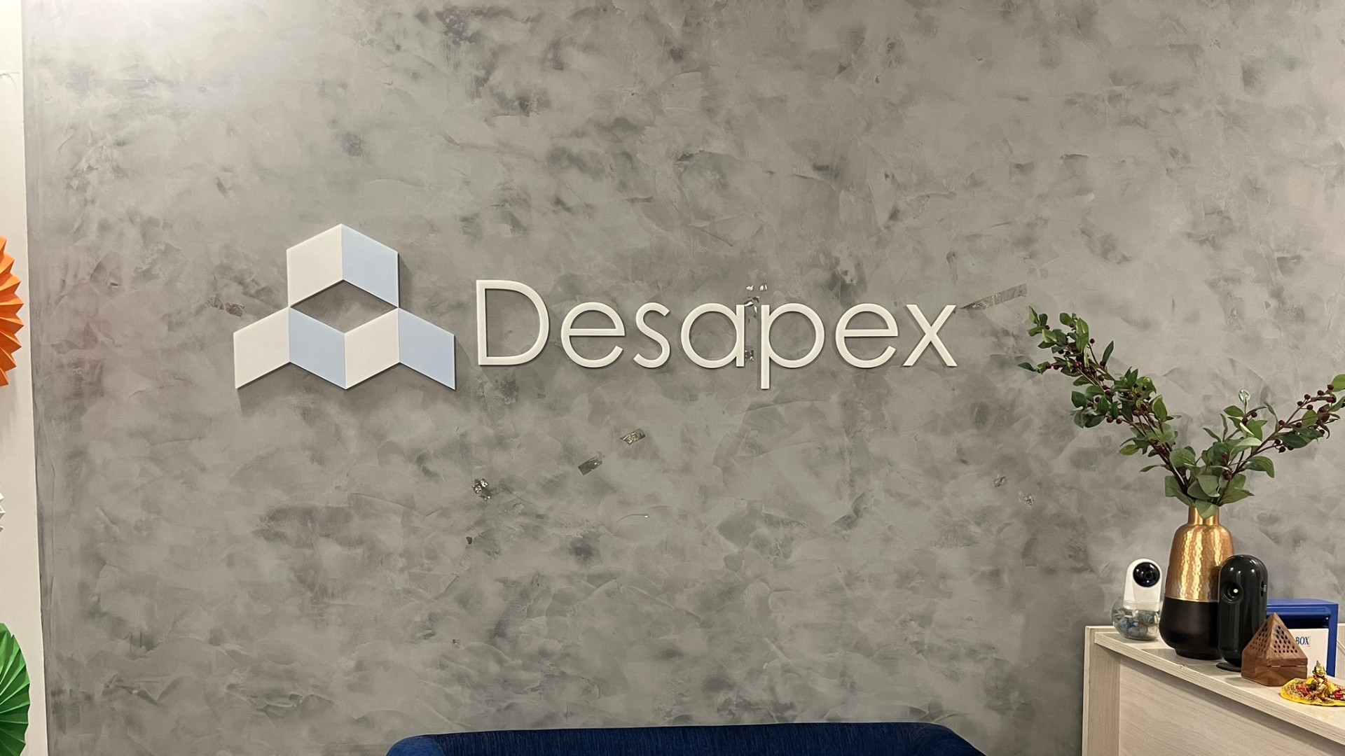 A man stands while two people sit at a table in an office setting, smiling and engaged in conversation. The mood is friendly and collaborative.