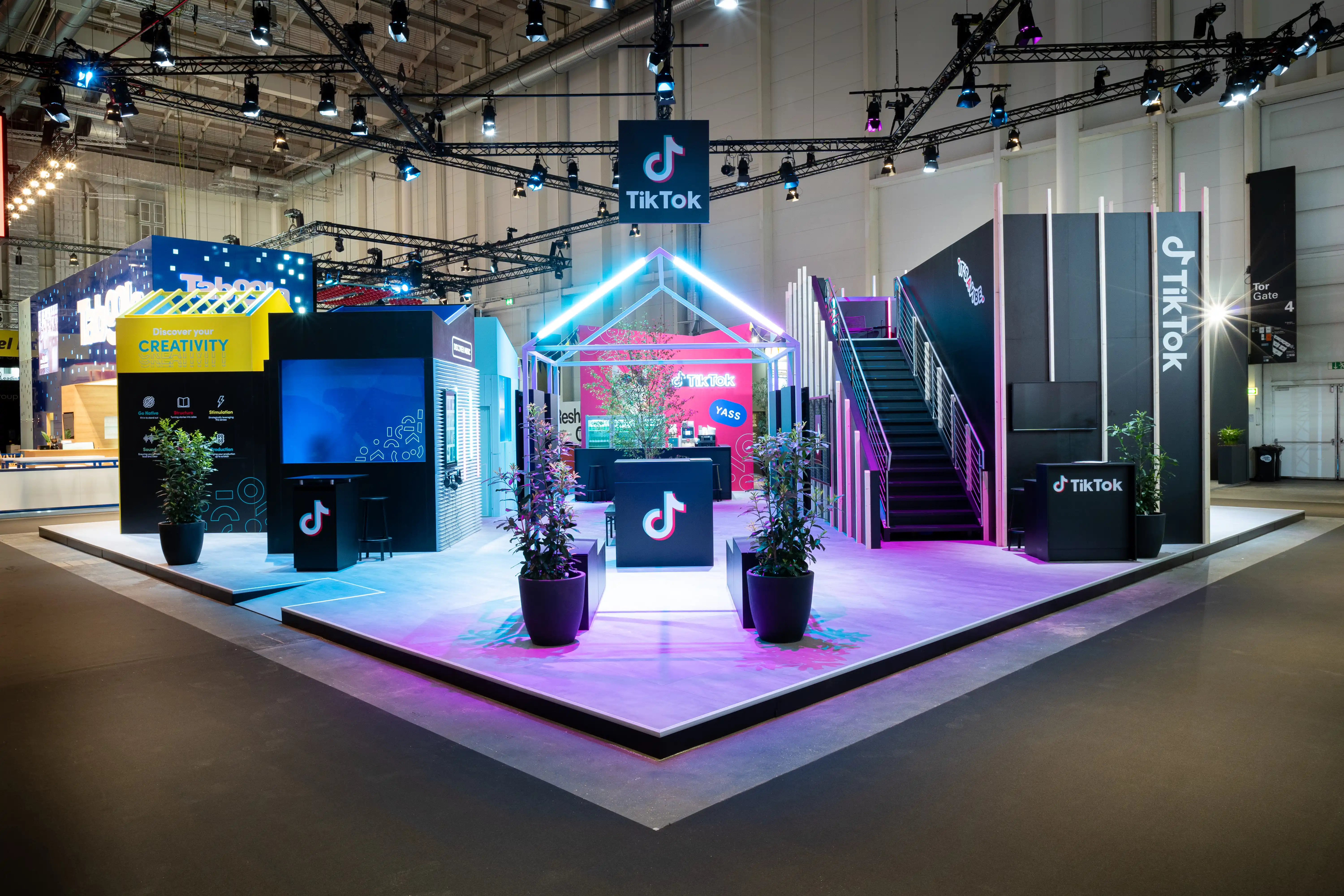 TikTok booth at OMR with neon house outline, black stair tower and pink-lit floor under the truss ceiling.