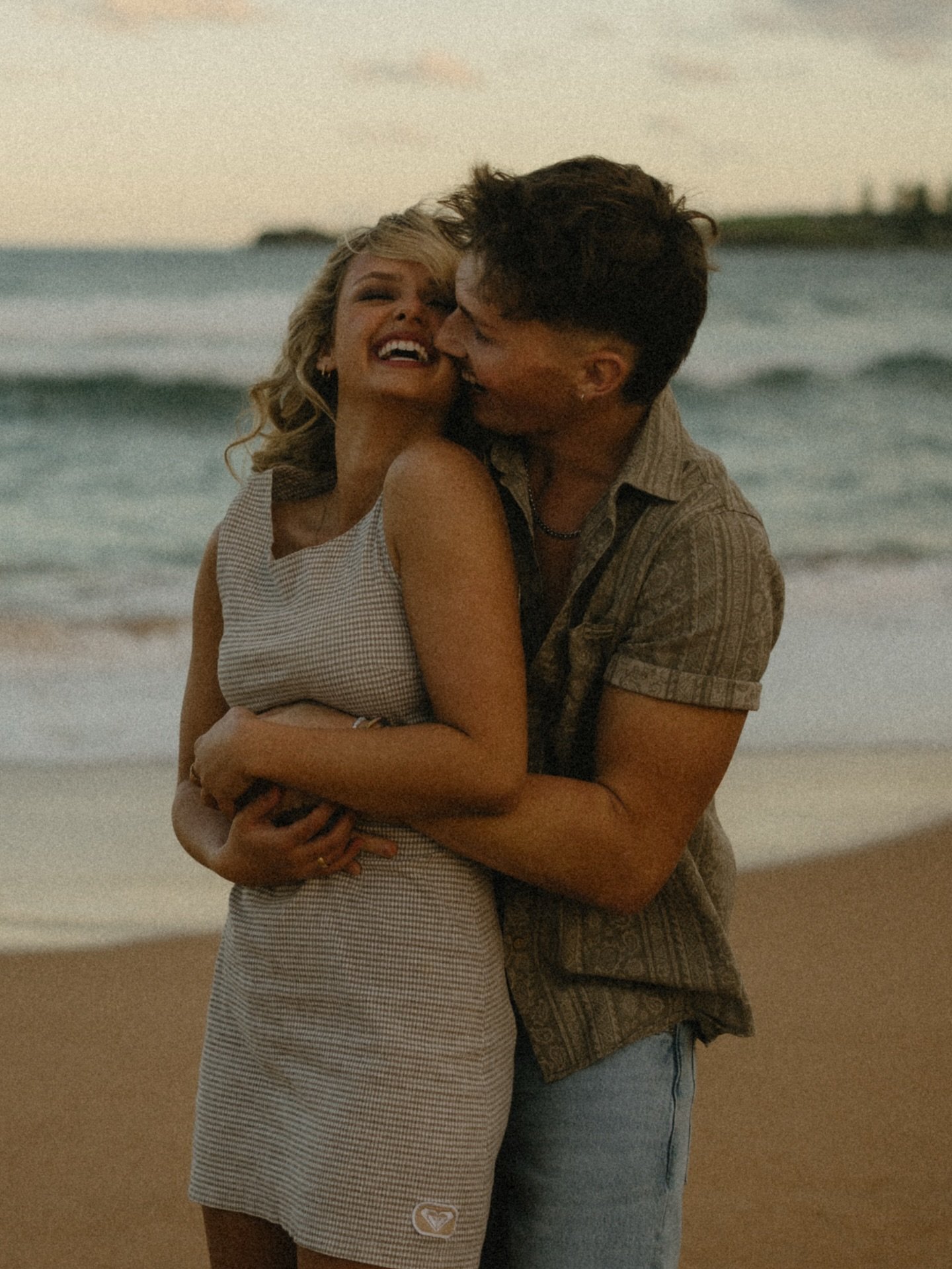 A joyful couple embraces and laughs on a sandy beach at sunset, with gentle waves in the background, creating a romantic and serene atmosphere.