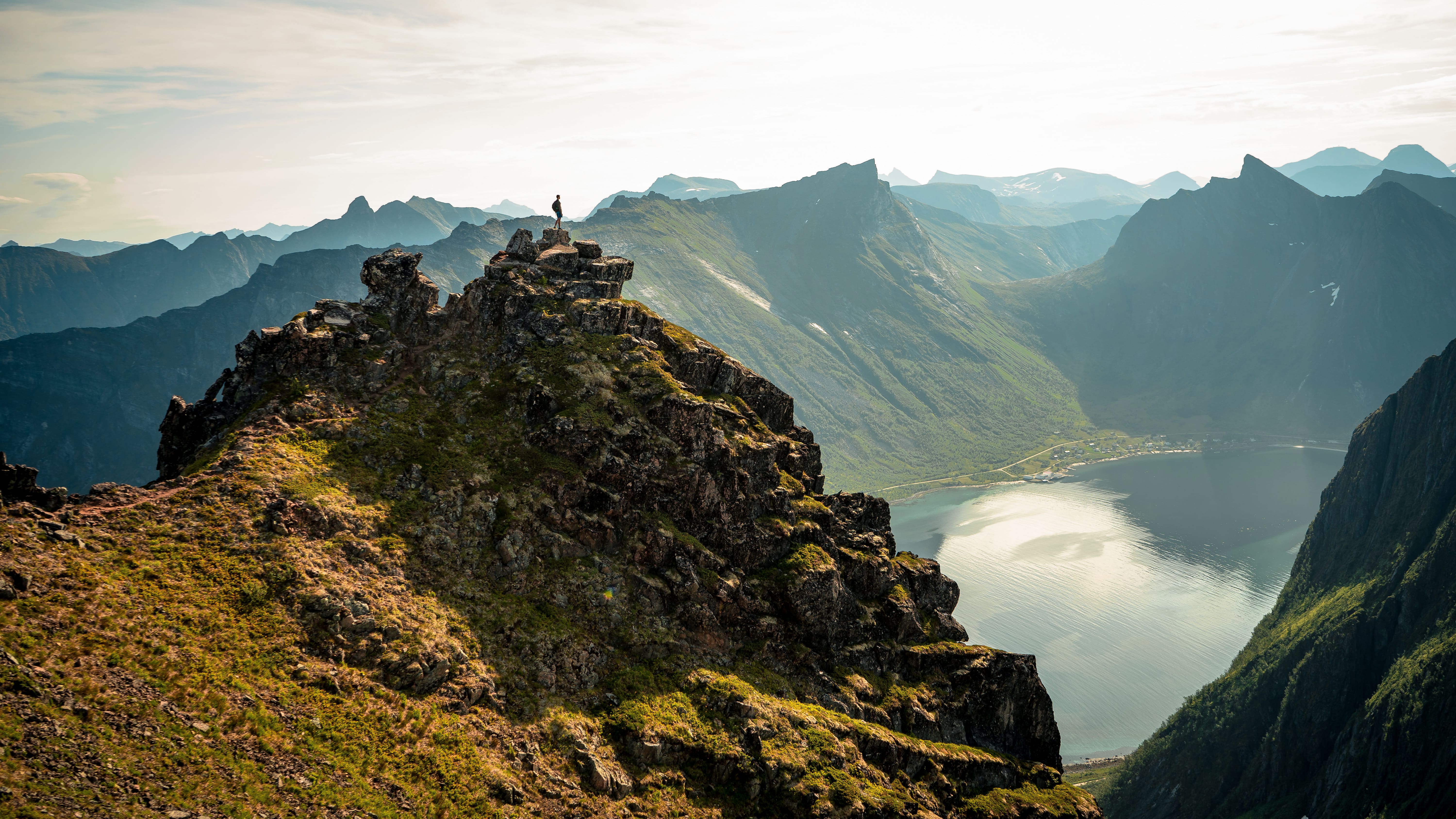A lone hiker stands atop a rugged mountain peak overlooking a serene fjord, surrounded by dramatic cliffs and expansive views under a partly cloudy sky.
