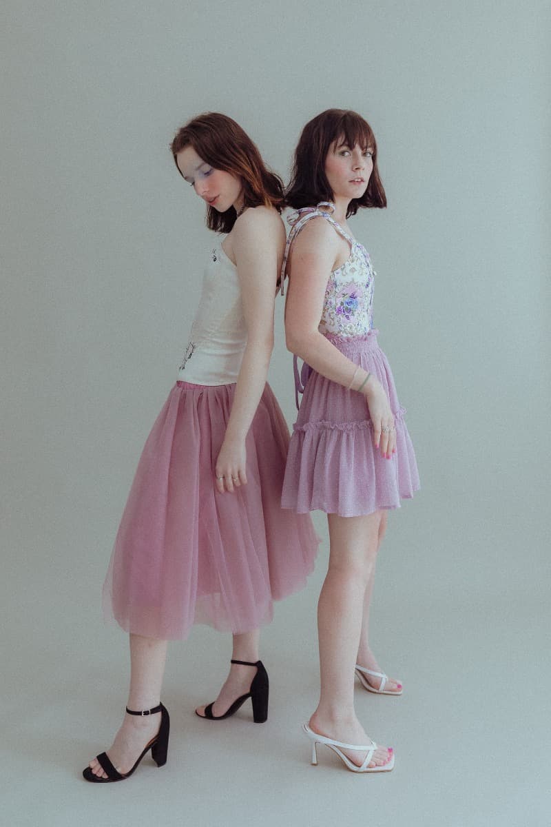 Two models standing back-to-back, one in a white top and pink tulle skirt, the other in a floral top and pink layered skirt, during an ethereal photoshoot at Revelator Studio in Shreveport.