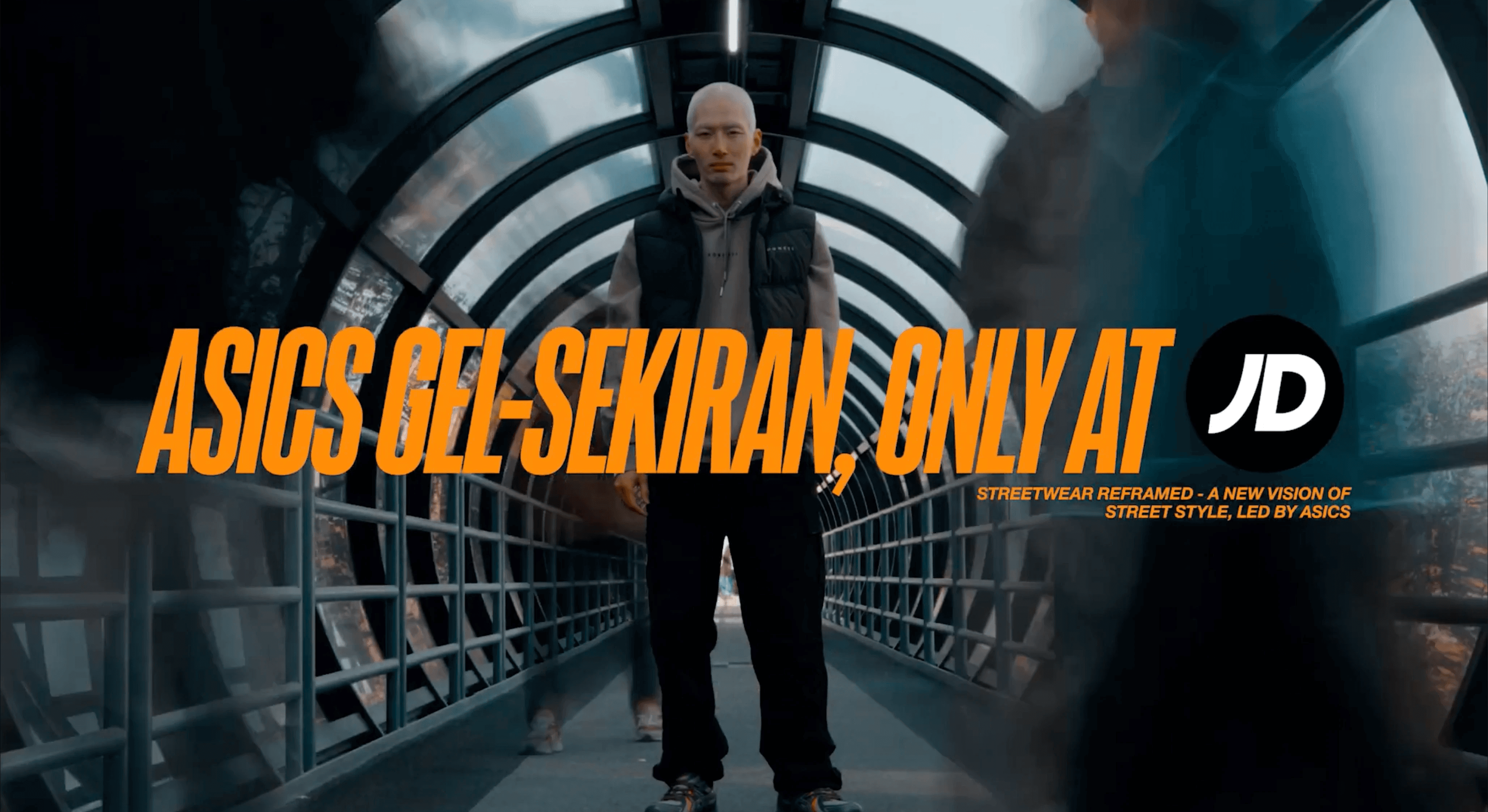 A person stands confidently on a futuristic, metal-arched bridge wearing athletic streetwear, showcasing the ASICS GEL-SEKIRAN shoes in a promotional image with bold text for JD Sports.