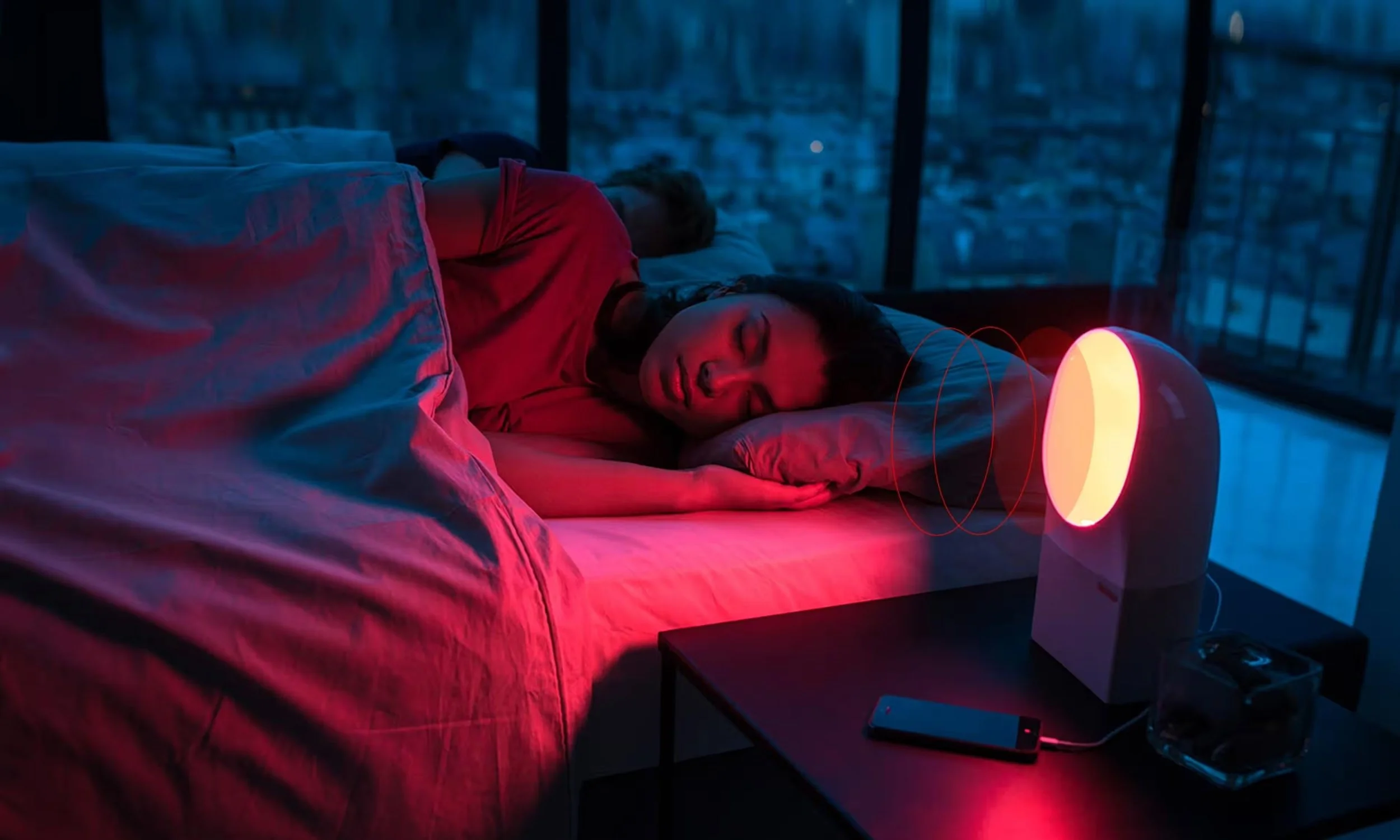 Two people sleeping in a dark room lit by a warm red bedside light device