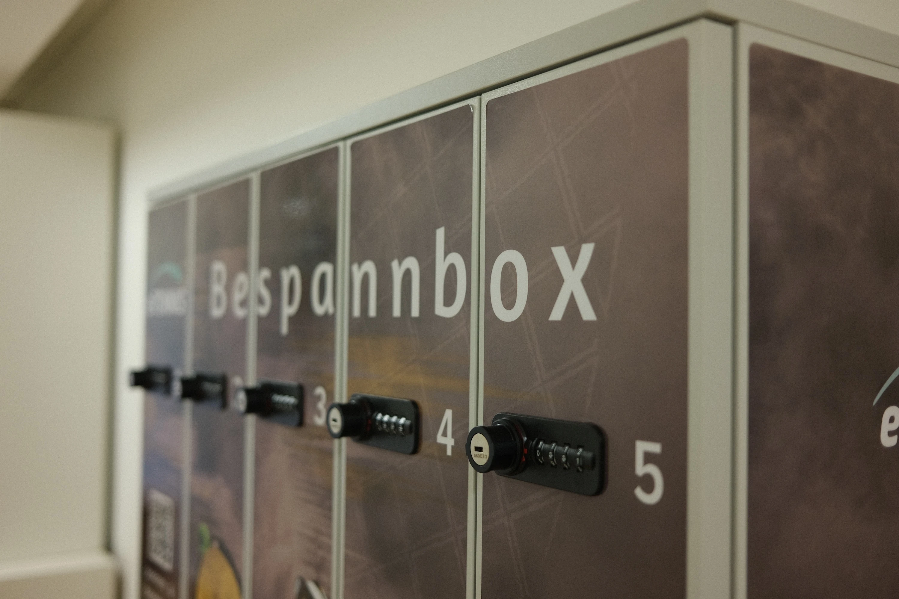 Eine Bespannbox mit fünf nummerierten Fächern, die jeweils mit einem Zahlenschloss gesichert sind. Die Box ist mit einem Tennis-Motiv bedruckt und trägt das Branding von “eTENNIS” und “Stefs Racket Corner”. Ein QR-Code auf der linken Seite weist darauf hin, dass Kunden ihre Schläger für eine Bespannung abgeben können.