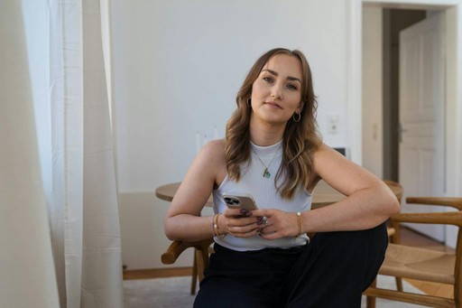 A woman with long hair sits casually on a chair, holding a smartphone and looking at the camera.