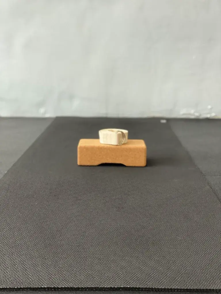 A silver ring with a geometric design is displayed on a small wooden stand against a dark fabric background.