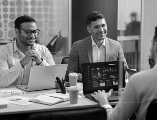 Group of professionals in a meeting room, smiling and discussing while working on laptops, with charts and graphs displayed on one screen.