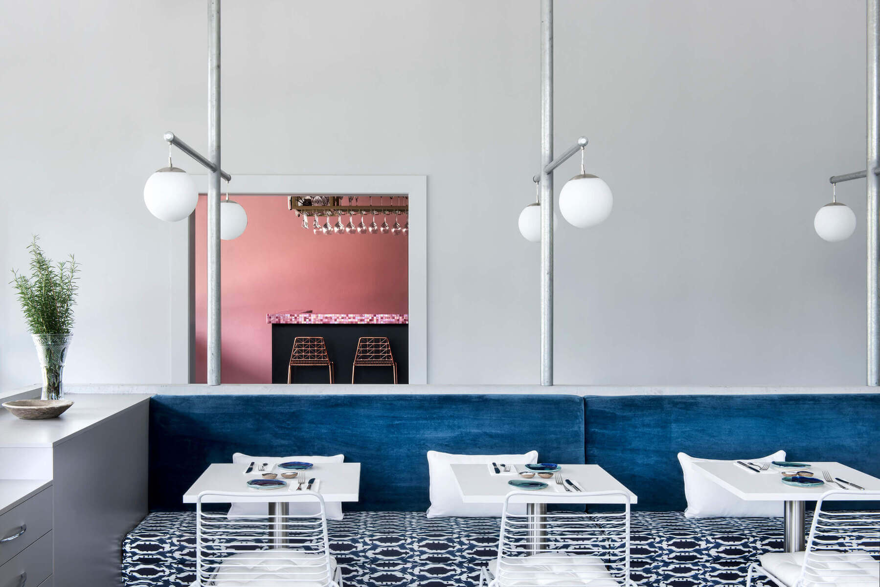 Contemporary restaurant dining room interior with blue banquette seating, white metal wire chairs, globe lights, and a doorway opening to a pink bar room.