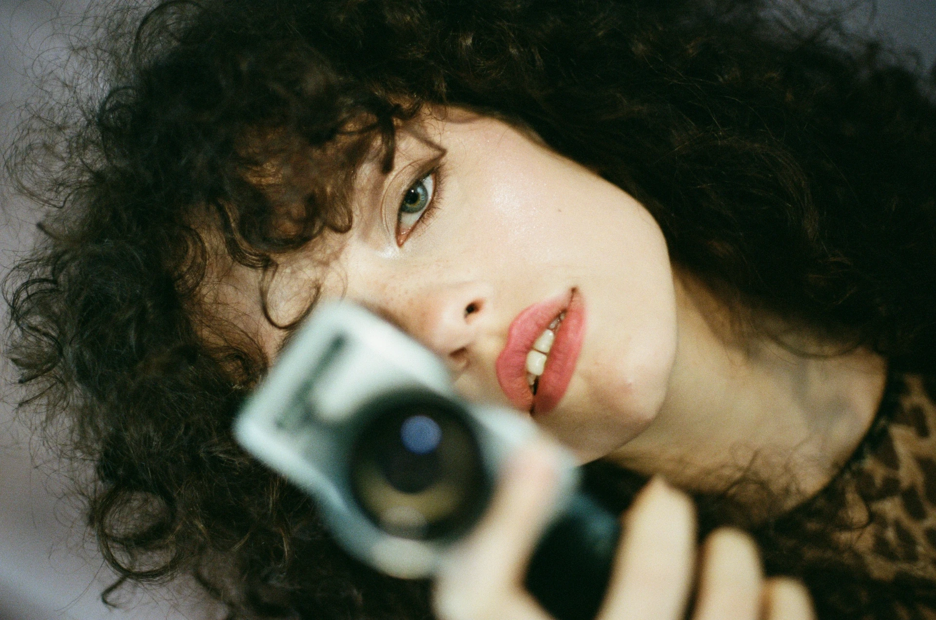 Schwarz-weißes Porträt einer Frau mit lockigem Haar, die direkt in die Kamera blickt und eine Kamera in der Hand hält; das Objektiv ist im Vordergrund unscharf.