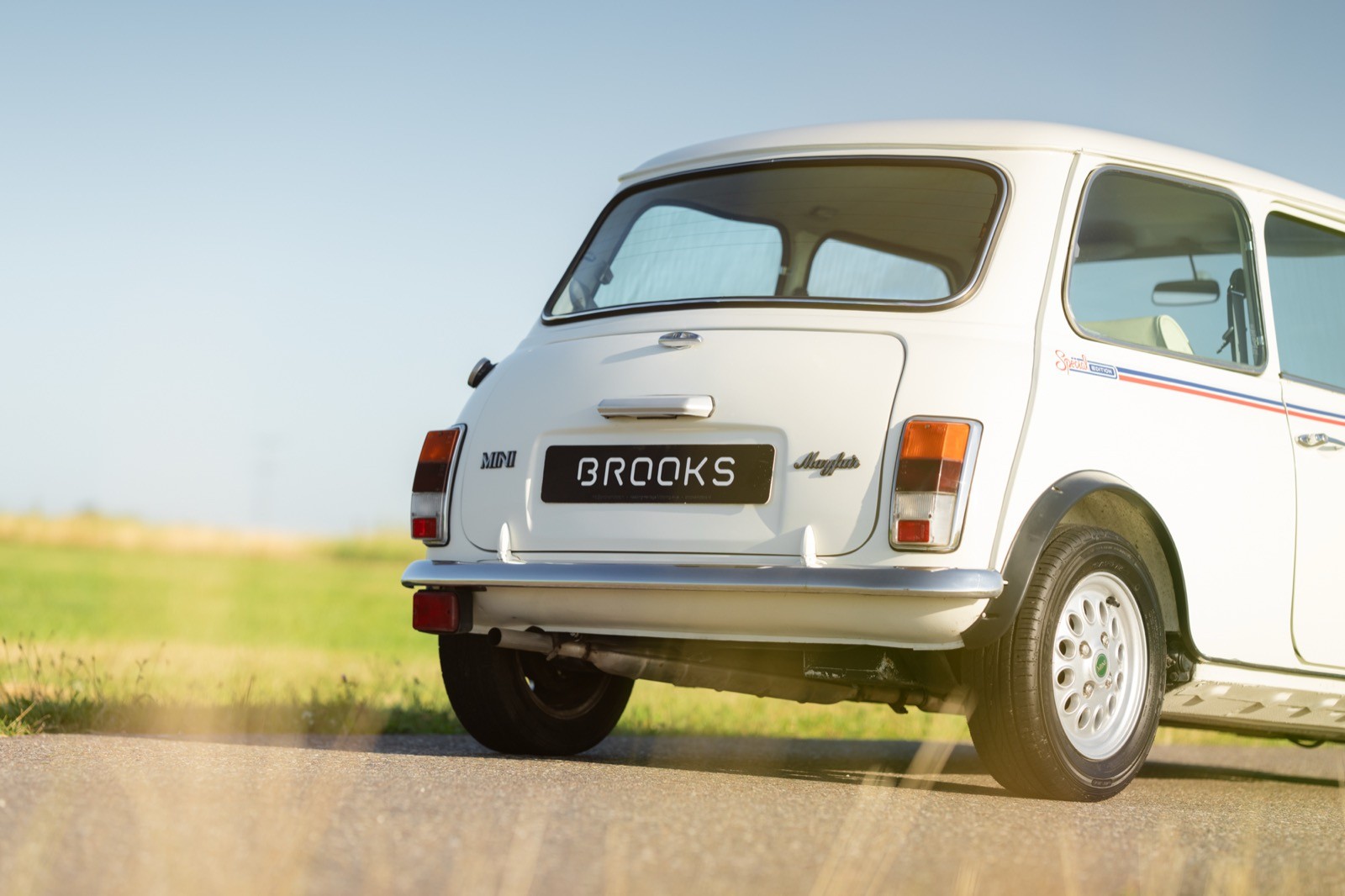 A vintage white car parked on a road with green fields in the background under a clear blue sky.