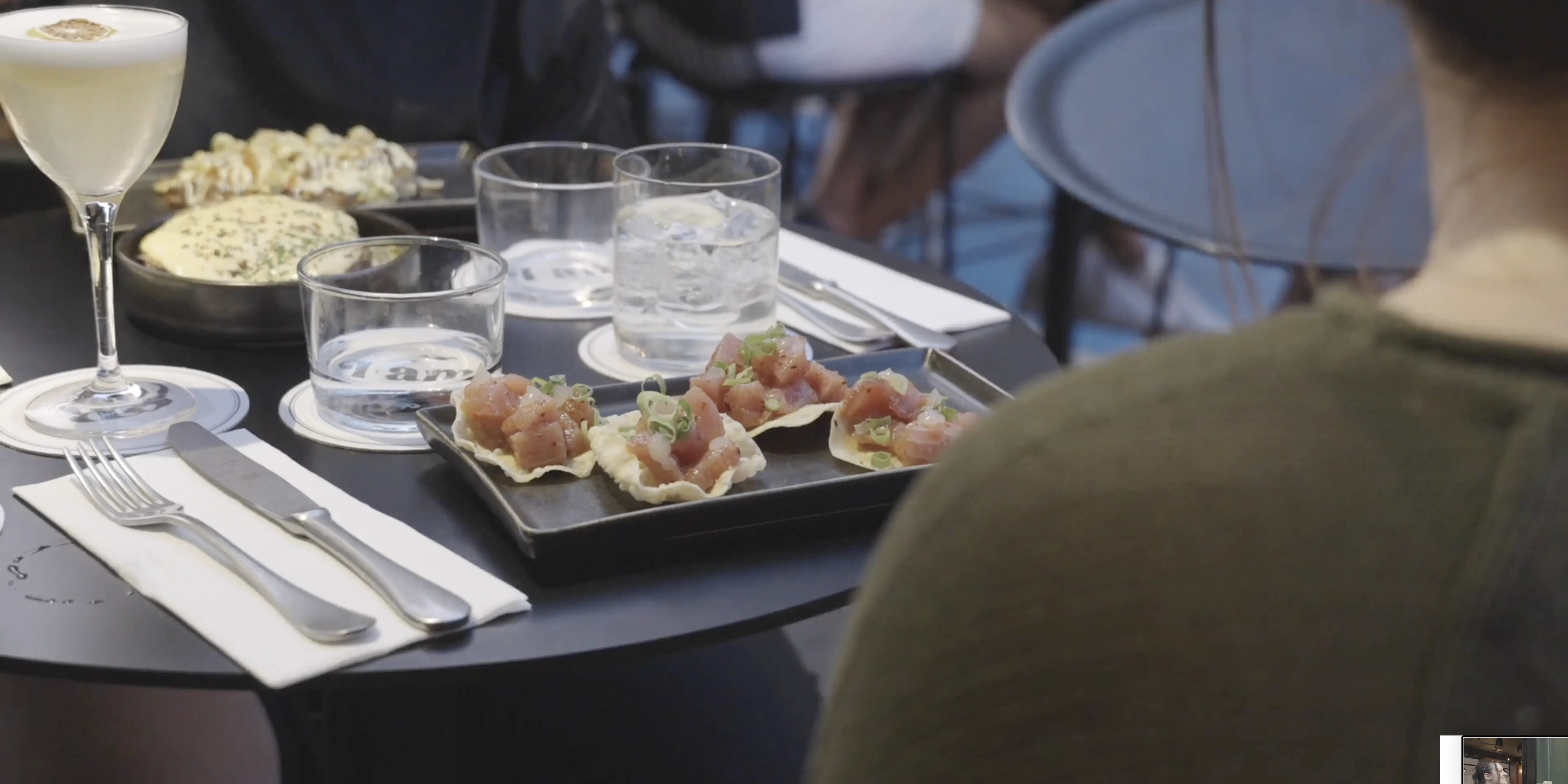 A black table set with plates of gourmet appetizers, including tuna tartare on crackers, a round serving of creamy cheese spread beside crunchy flatbreads, and two glasses of clear cocktails, emphasizing a fine dining experience.