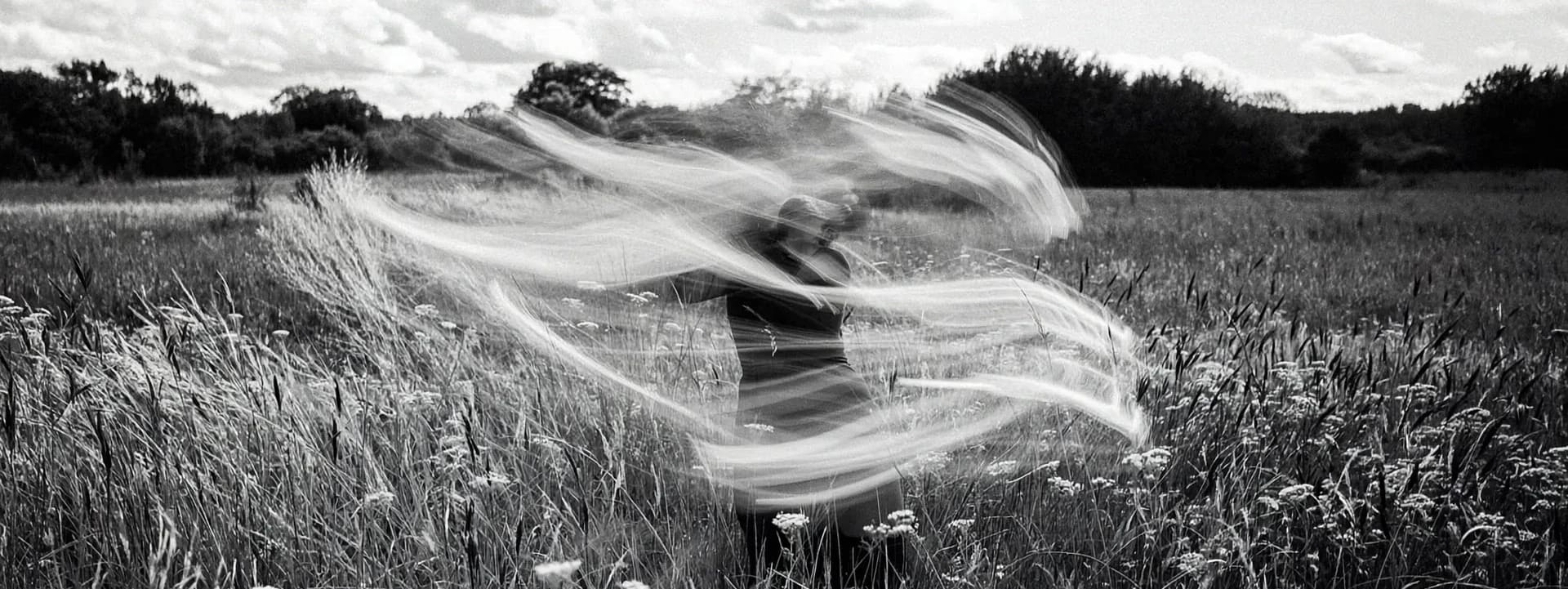 Black-and-white photograph of a person in motion, creating a blurred effect, standing in a field of tall grass and wildflowers under a cloudy sky, capturing a sense of freedom and nature."