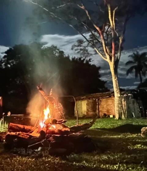 Uma fogueira brilhante crepita sob a lua cheia, com uma árvore recortada contra o céu noturno e as nuvens.