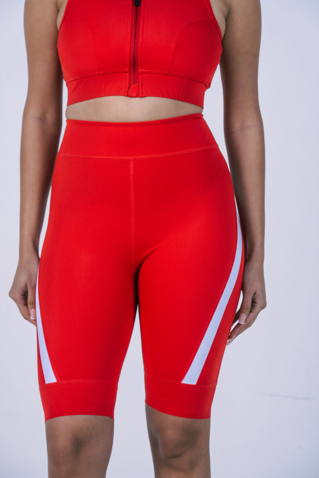 A-close-up-studio-shot-focusing-on- the-midsection-and-legs-of-a- woman-wearing-bright-red-high-waisted-cycling-shorts-The-shorts- feature-a-white-graphic-stripe-that- curves-diagonally-up-from-the-hem -on-both-outer-sides-of-the-legs- The-model-is-also-wearing-a- matching-red-sports-bra-crop-top- with-a-visible-front-zipper