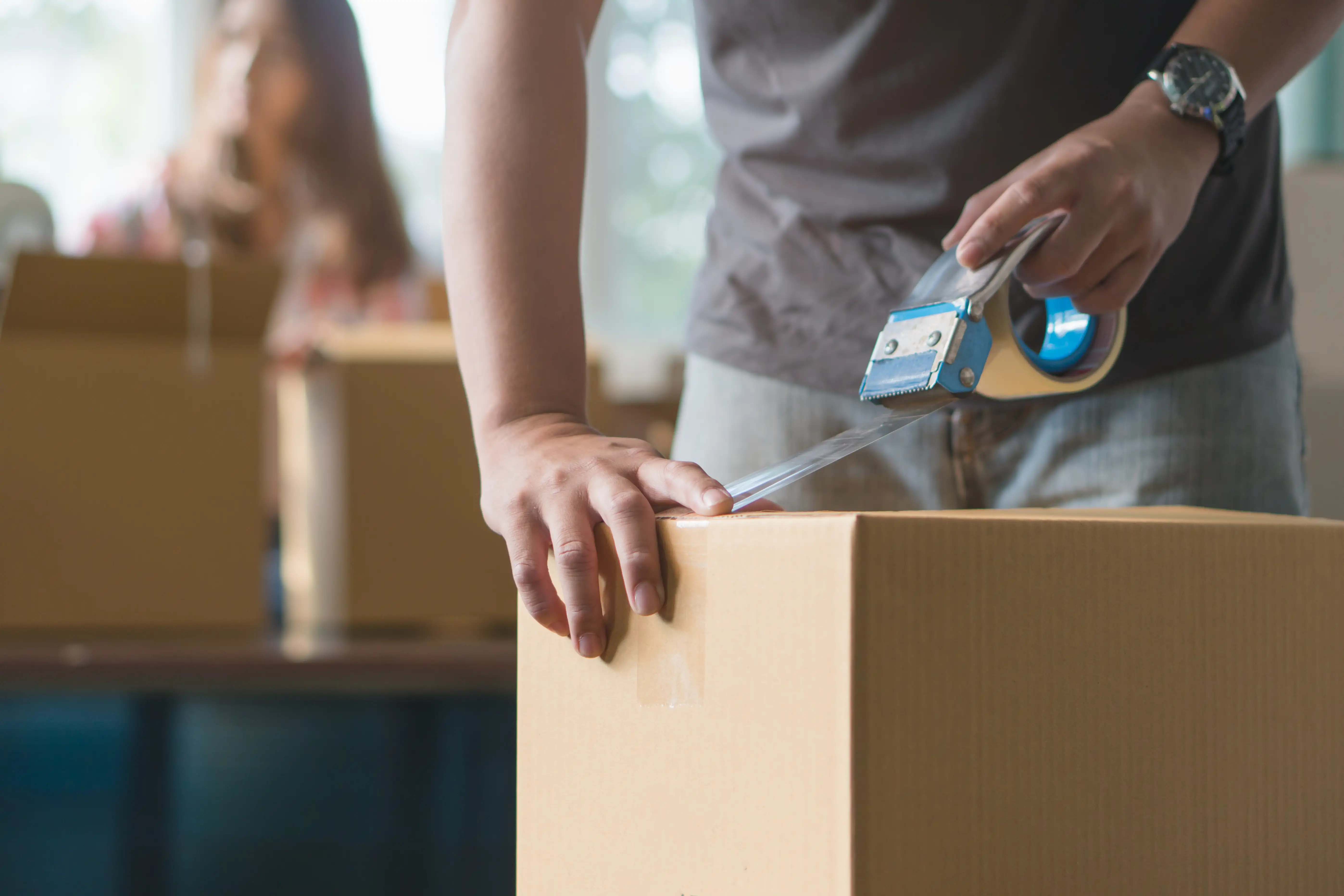Personal sealing a cardboard box with a tape dispenser while packing for a move, illustrating intra-state residence changes under California Labor Code Section 970