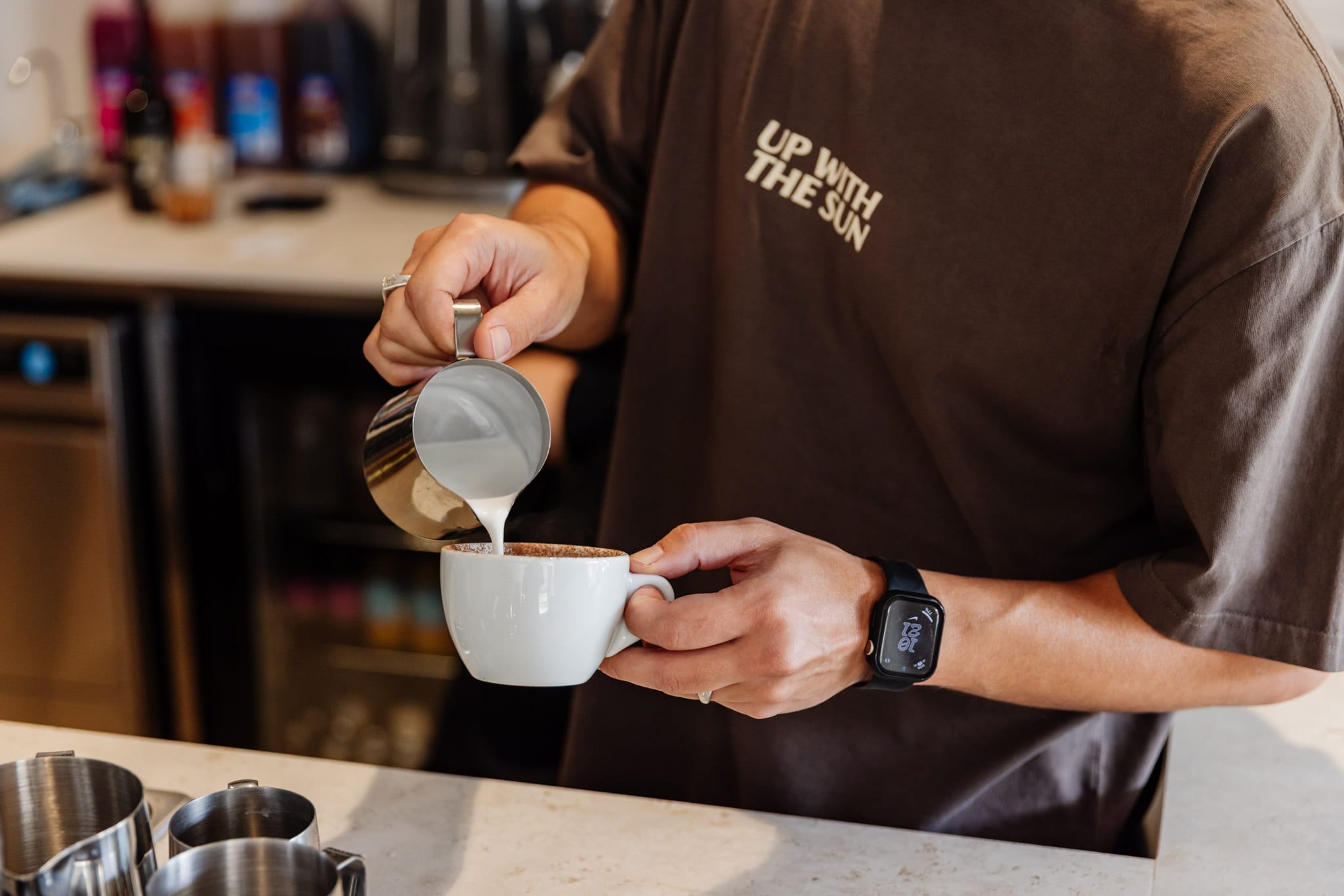 Person wearing a beige apron whisking green matcha tea in a ceramic bowl on a kitchen counter.