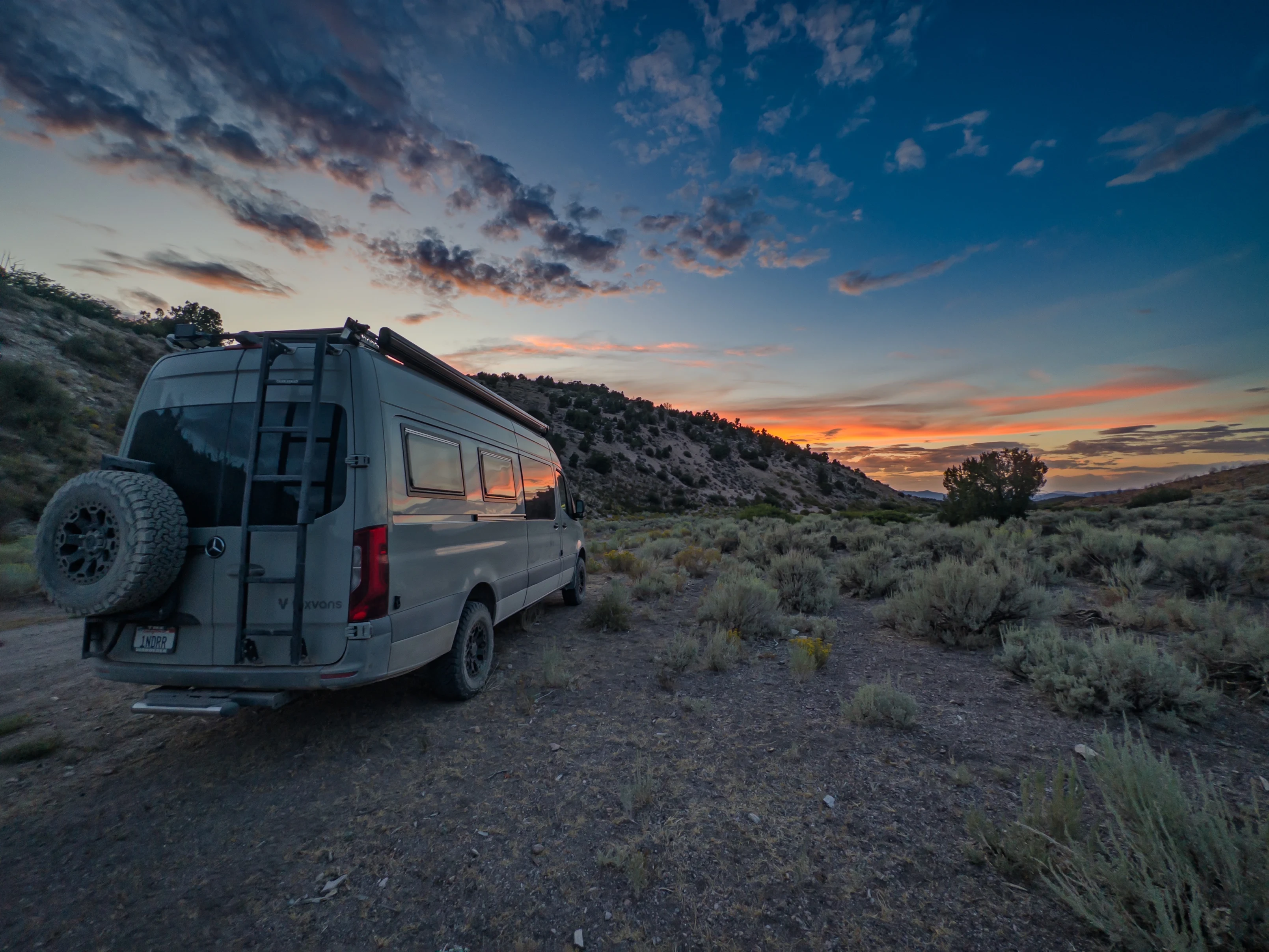 A scenic view of a camper parked on rocky terrain under a colorful sunset sky.