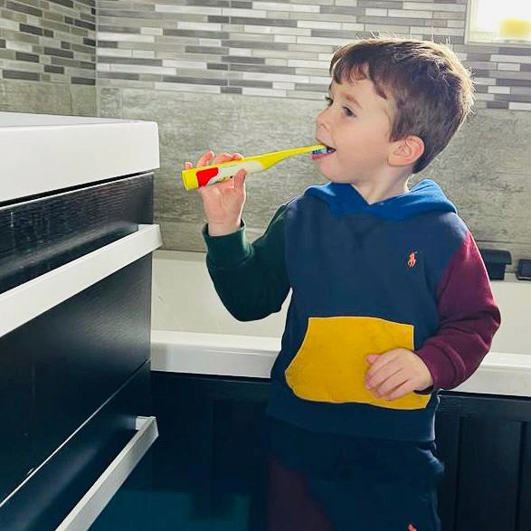 A young child brushes their teeth in a bathroom. They're stood next to the sink holding an electric toothbrush.