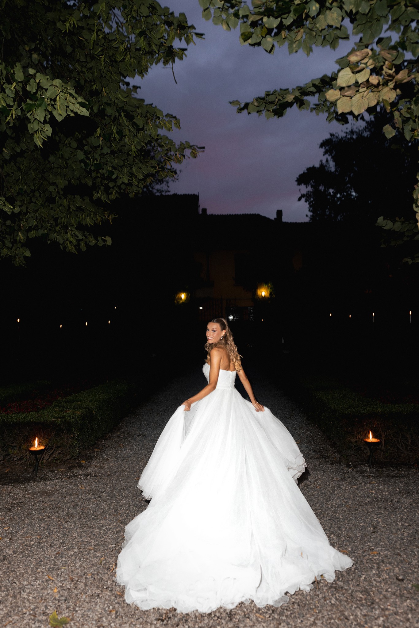 Sposa in abito bianco cammina al tramonto nel parco di Villa Ferrari tra luci notturne durante il matrimonio
