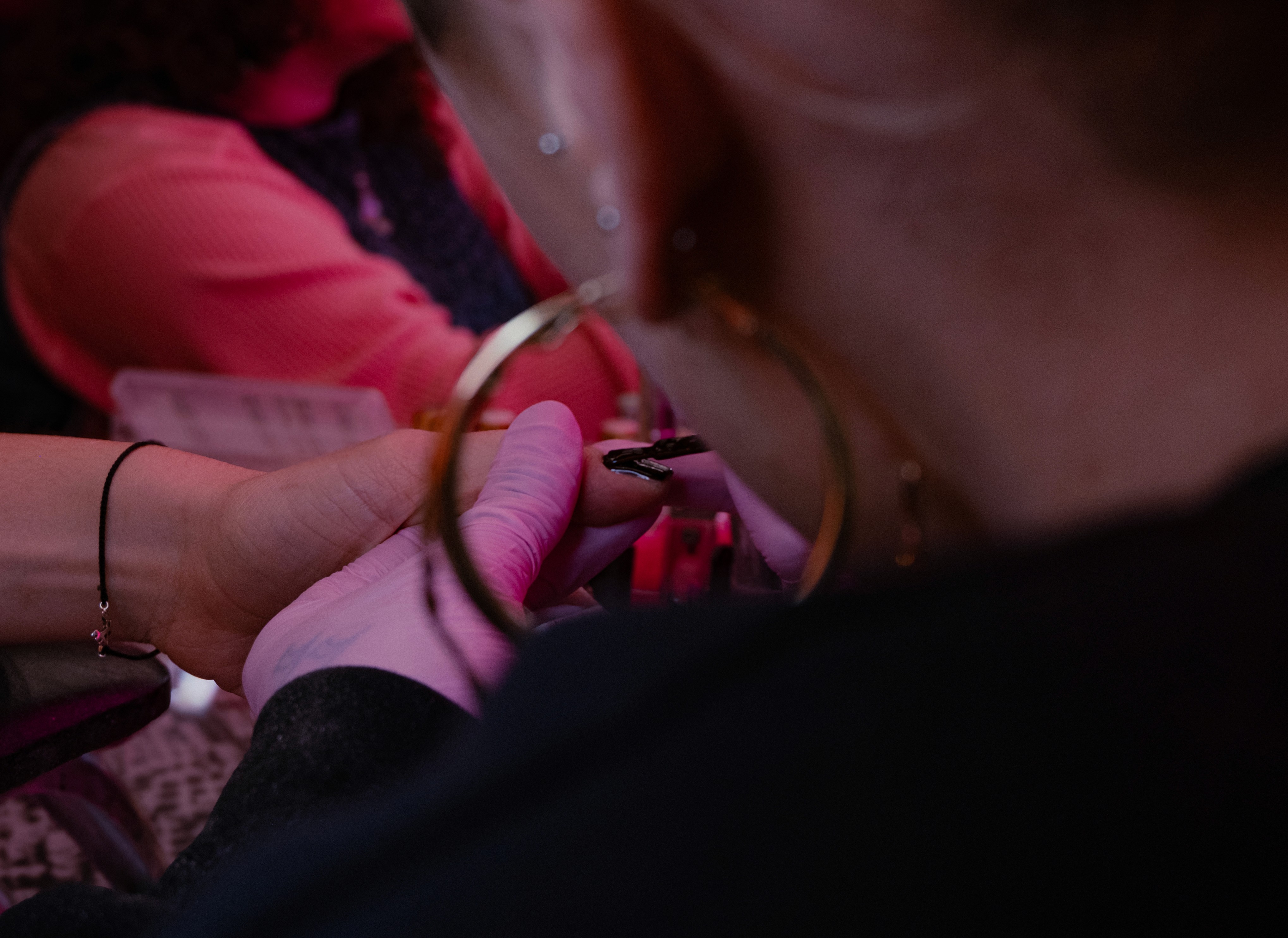 A focused shot over the shoulder of a technician (wearing large gold hoop earrings) as they work on a client’s nail under pink lighting.
