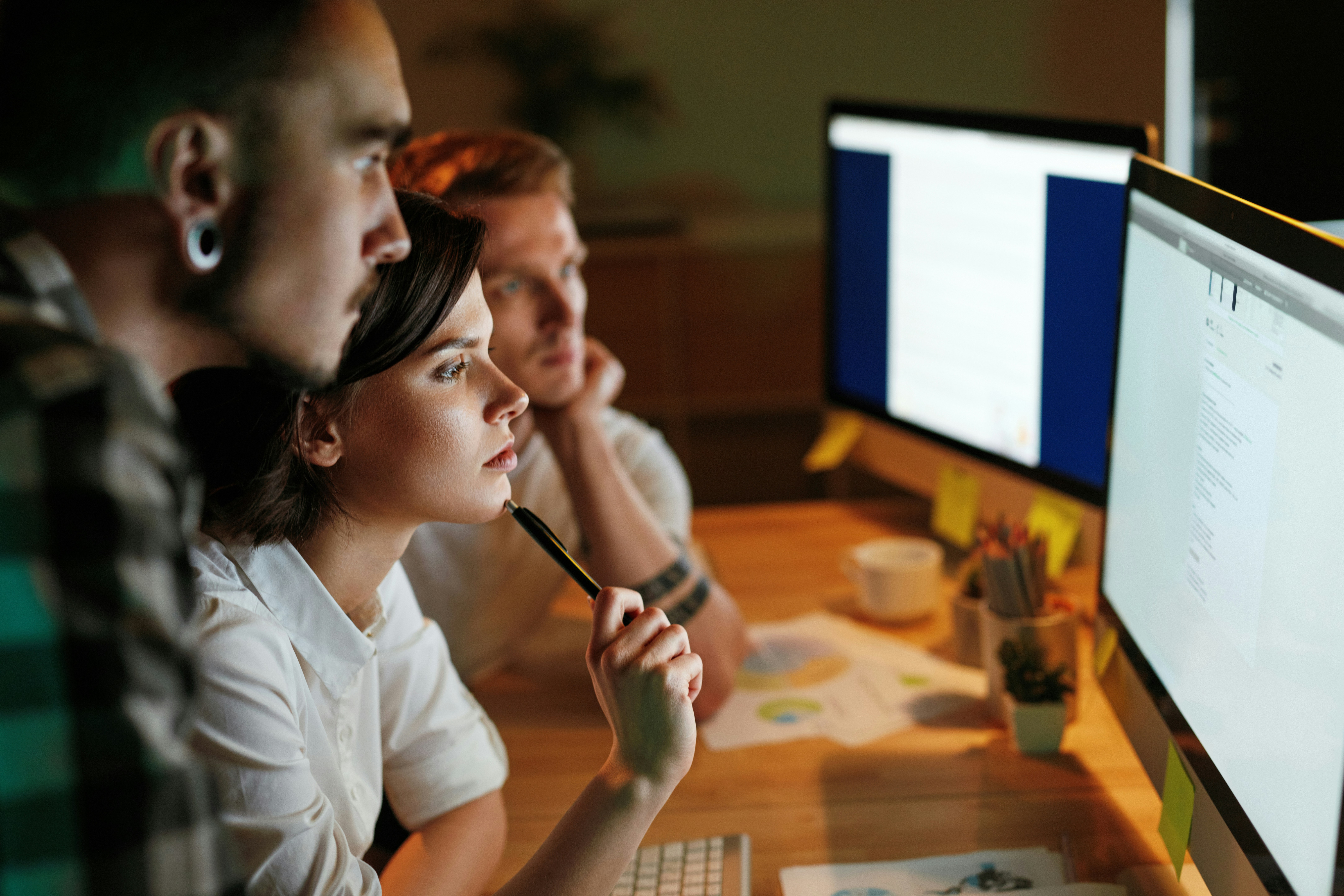 Three individuals focus on computer screens, discussing their work in a dimly lit setting.