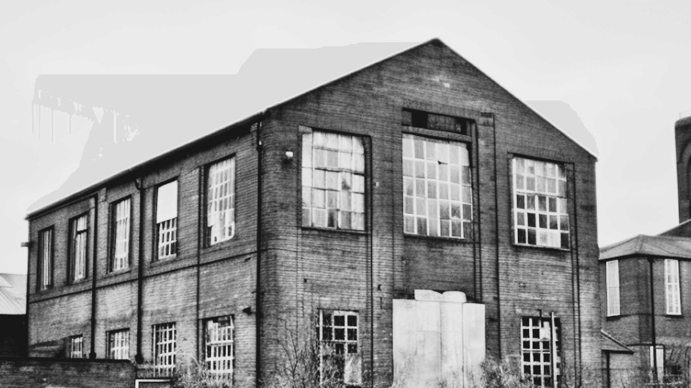 A black and white photo of a brick building with large windows, featuring a symmetrical design under a cloudy sky.