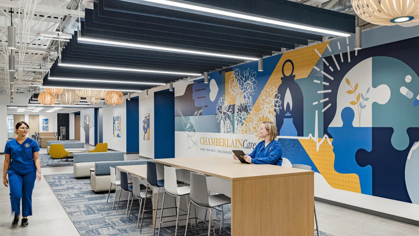 A modern academic lobby with blue and white decor. A seated person at a wooden table interacts with a tablet. Another person in scrubs walks past. A colorful mural depicting education themes is on the wall, emitting a welcoming and scholarly atmosphere.