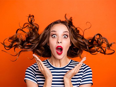 A surprised woman with wavy brown hair is against a vibrant orange background, wearing a black and white striped shirt, with her hands near her face and mouth open in amazement.