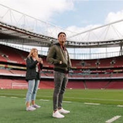 Due persone sono in piedi su un campo da calcio all'interno di un grande stadio con tribune rosse e una vista parziale della struttura del tetto.