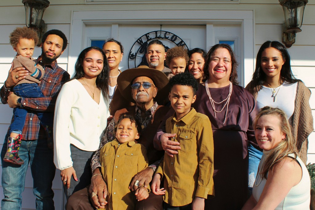 A diverse, multi-generational family gathered in front of a home, smiling warmly at the camera, representing family unity and togetherness across different ages and backgrounds.