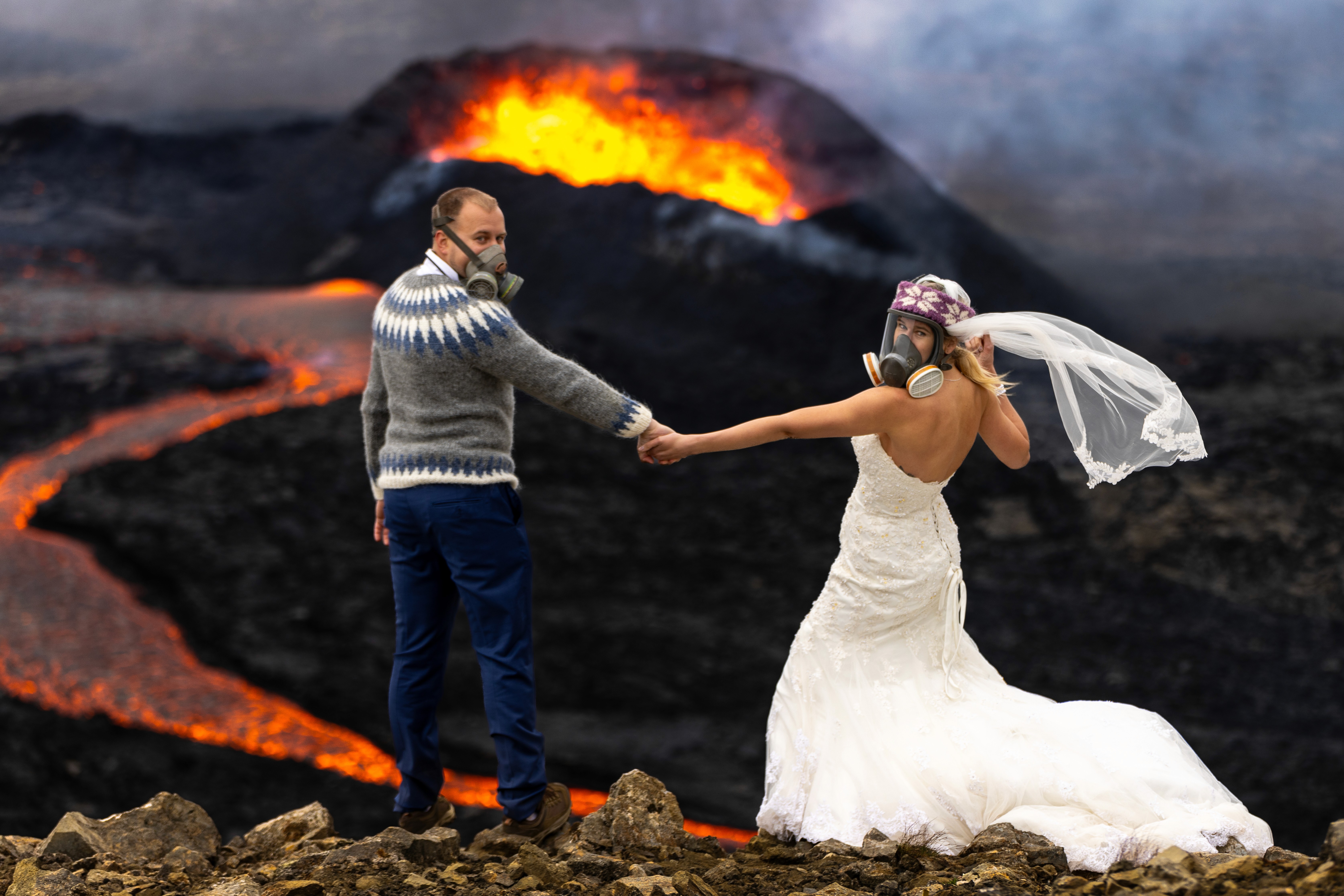 A bride and groom in formal wedding attire wear gas masks while holding hands in front of an erupting volcano, with flowing lava behind them.