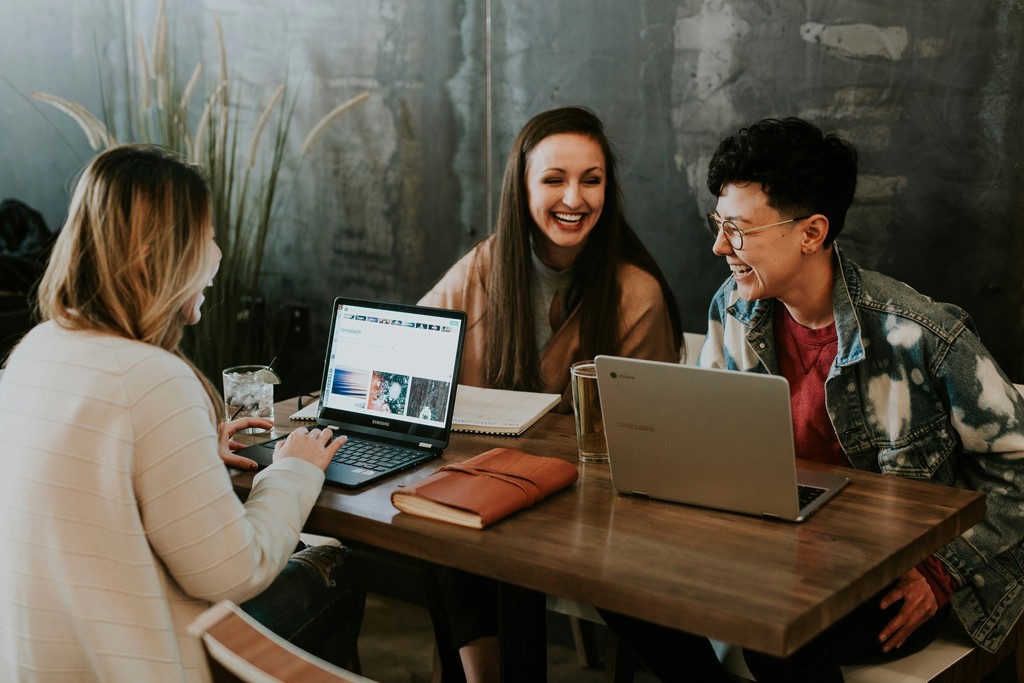 Three friends enjoying a collaborative work session at a trendy cafe, engaging with their laptops and sharing laughter around a wooden table.