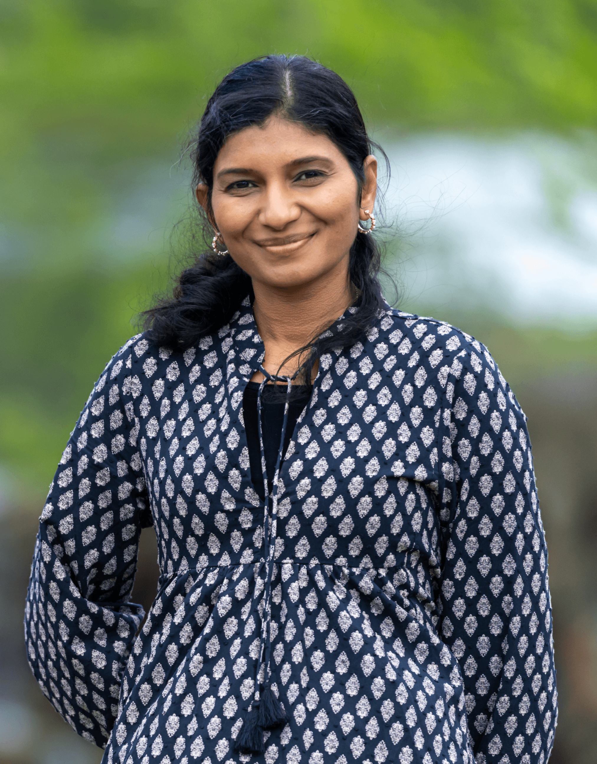 A woman with long dark hair, wearing a patterned black and white tunic, smiles warmly outdoors against a blurred natural background, illustrating a serene and welcoming aura suitable for an Ayurveda consultation.