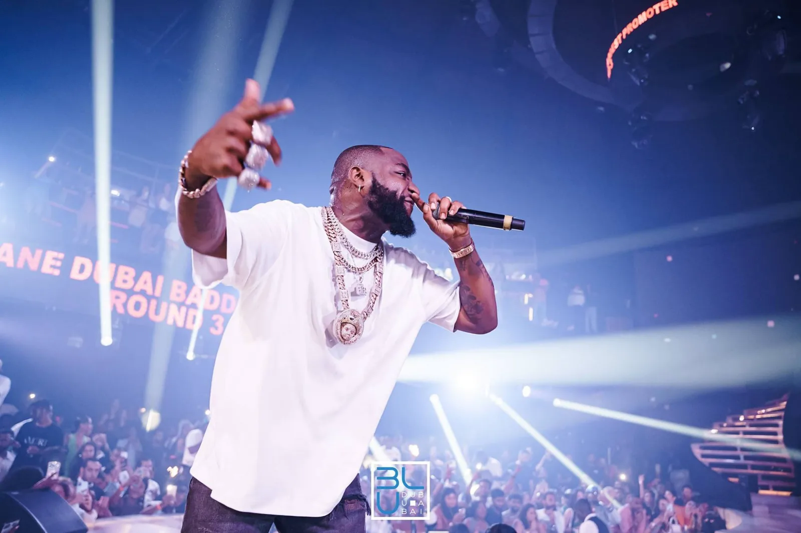 Artist in a white T-shirt rapping on stage at BLU Dubai, with light beams over a packed room