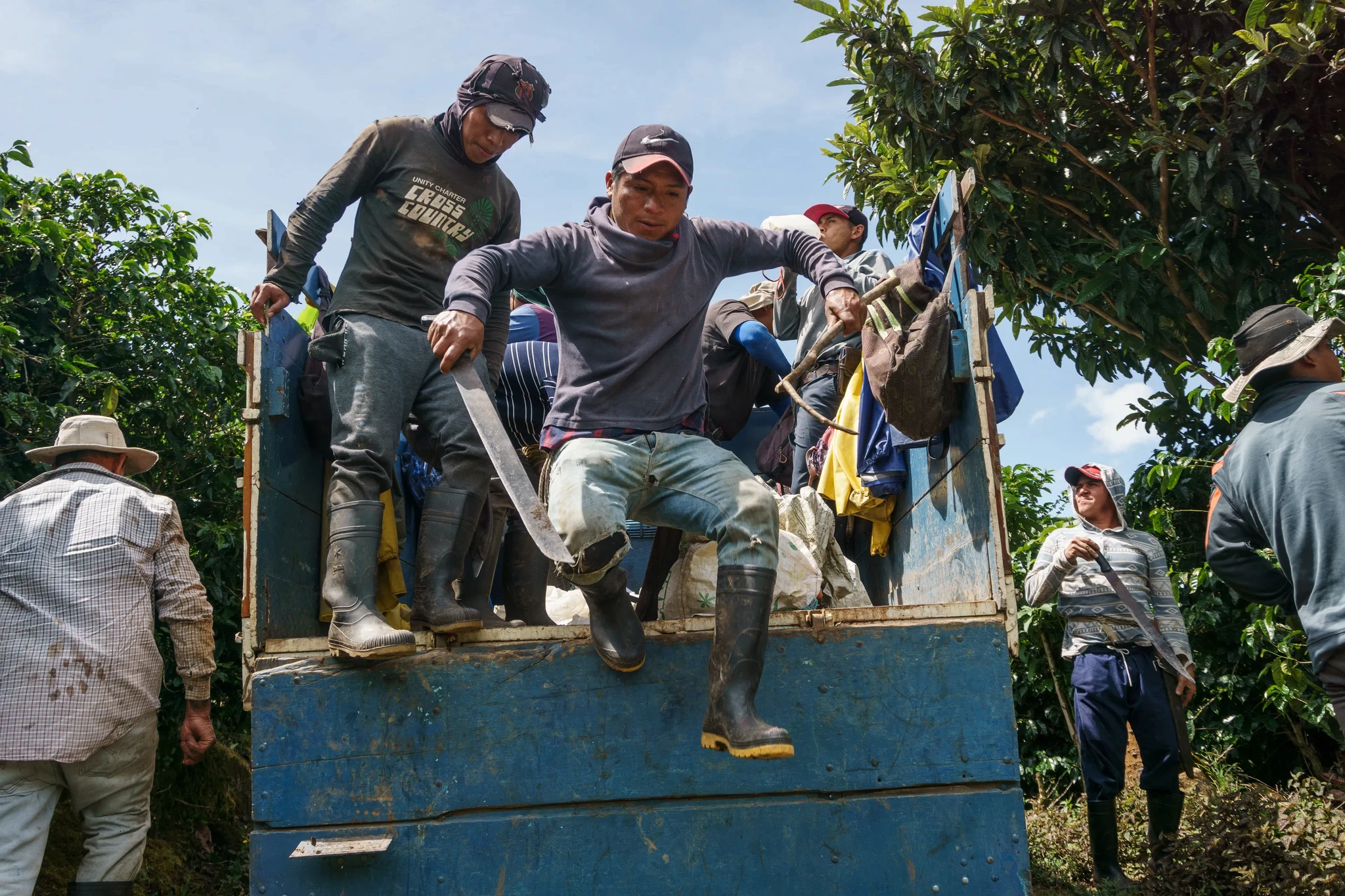 Workers, wearing hats and boots, unload from the back of a blue truck in a rural setting with trees and clear skies. One person is jumping down, holding a large machete. Others gather around or adjust their gear.