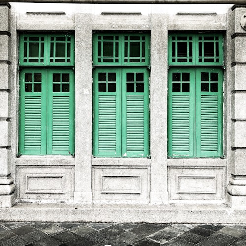 Facade with three green, shuttered windows set in a gray stone wall, featuring decorative rectangular frames beneath each window.