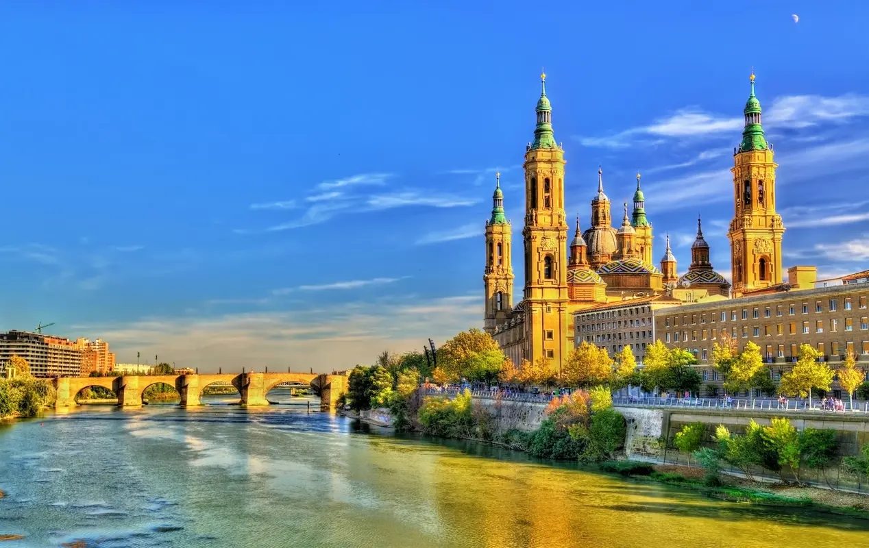 View of Zaragoza's Basilica del Pilar at sunset with reflections in the Ebro River, showcasing the city's iconic architecture and natural beauty.