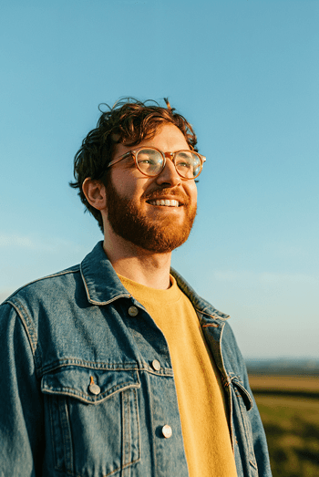 a young man with brown hair and beard wearing a glass, a yellow t-shirt under a denim jacket