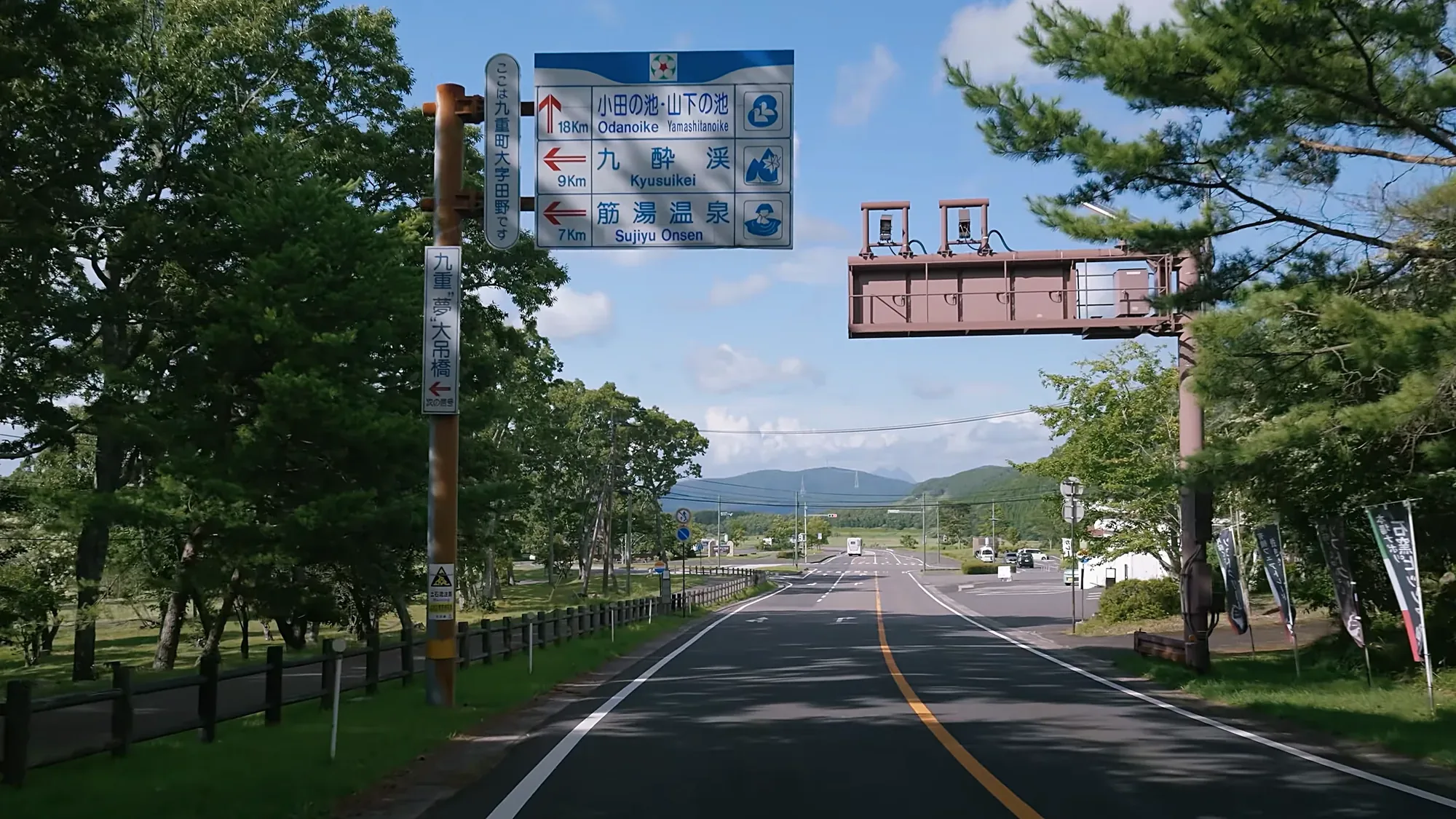 An approach road near Kurokawa Onsen with an overhead blue direction gantry and banner flags