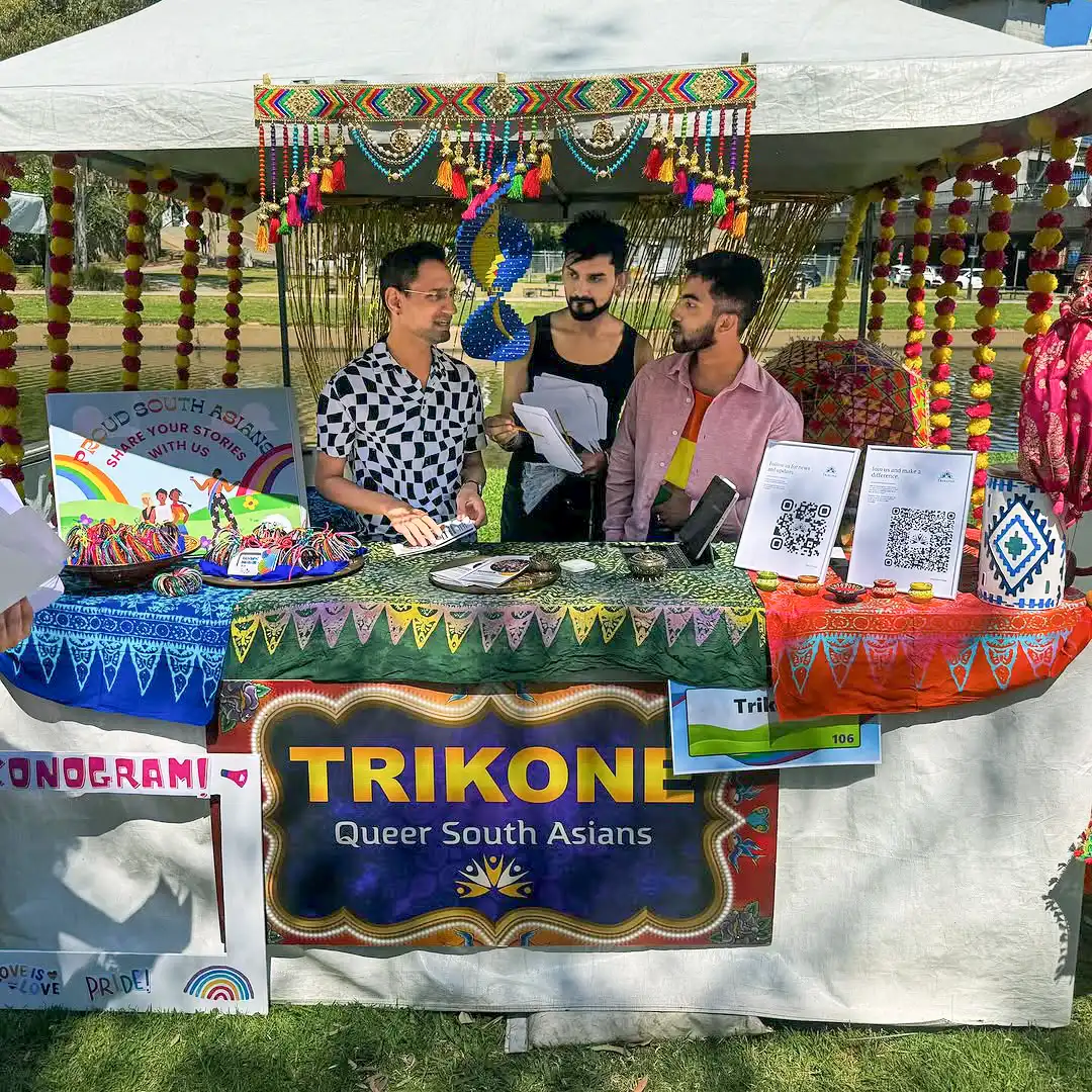 Three people stand at a colourful stall decorated with South Asian fabrics and rainbow accents. The stall sign reads "TRIKONE: Queer South Asians." QR codes, flyers, and art are displayed on the table.