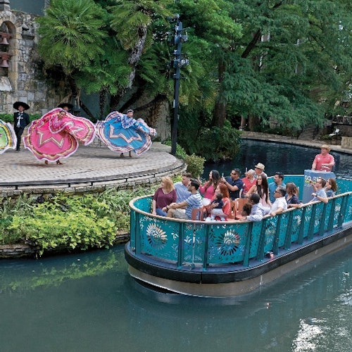 A boat of tourists watches dancers in colorful dresses perform on a riverside stage, surrounded by lush greenery.