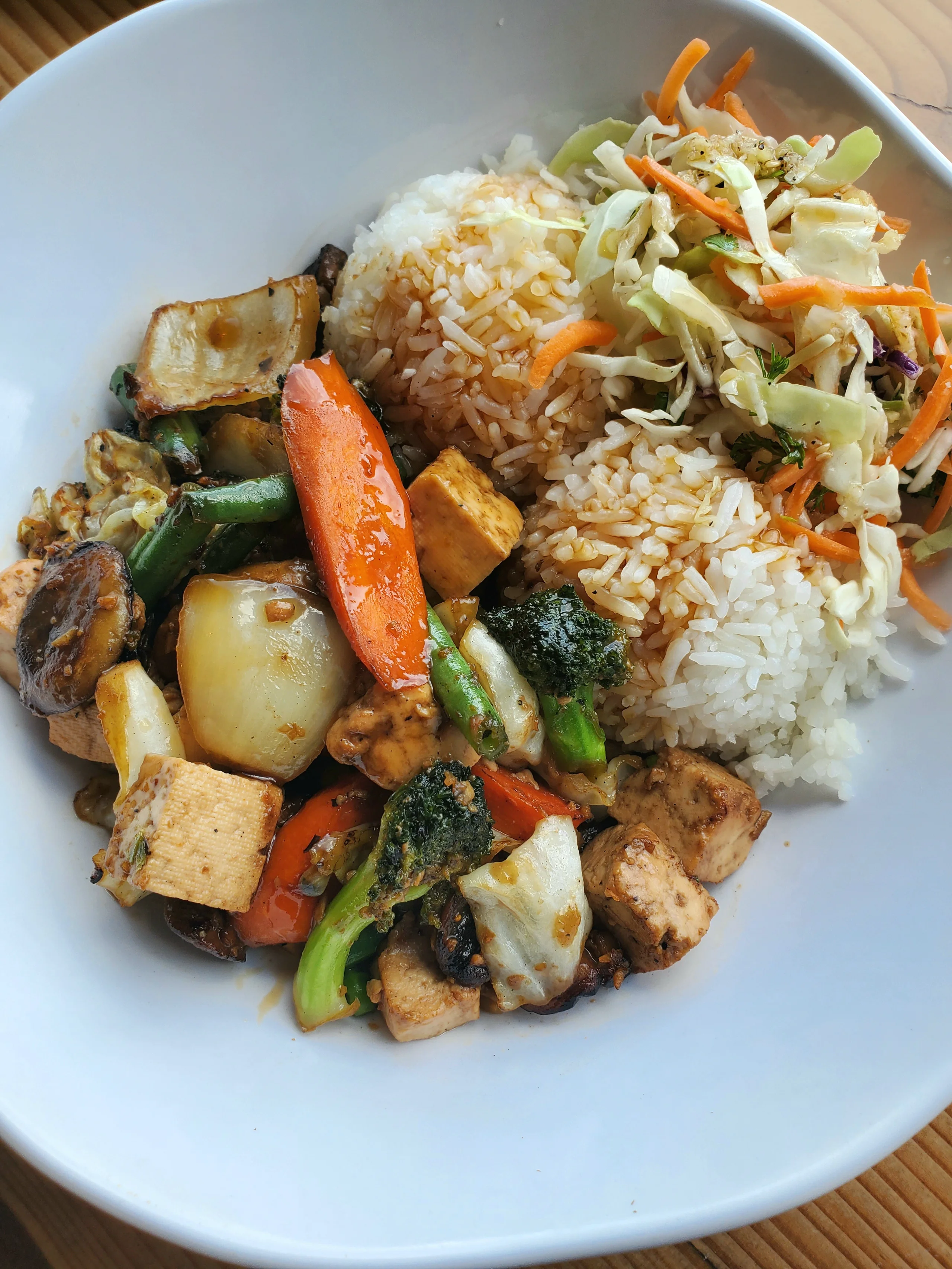 A white bowl filled with a colorful stir-fry of tofu, broccoli, carrots, and green beans in a dark ginger teriyaki sauce, served with jasmine rice and Asian slaw.