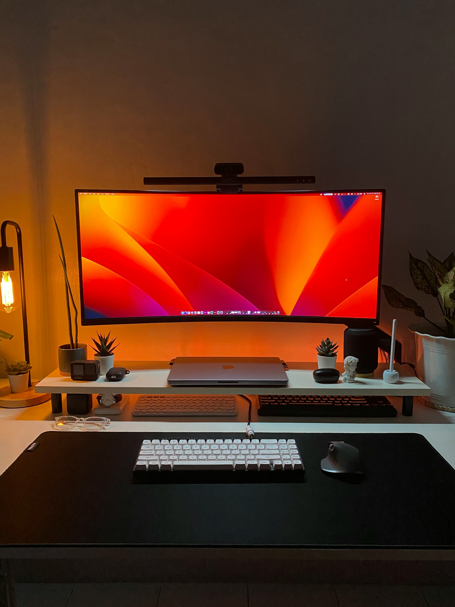 Dark desk setup with an ultra-wide monitor displaying an orange and red wallpaper, illuminated by a monitor light bar.