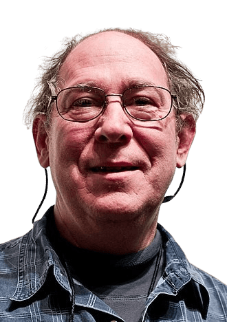 Color portrait of a middle-aged man with thinning hair, glasses, and a friendly expression. He is dressed casually in a plaid shirt over a dark undershirt, with eyeglass retainers hanging from his frames. His relaxed smile and approachable demeanor convey warmth and openness.