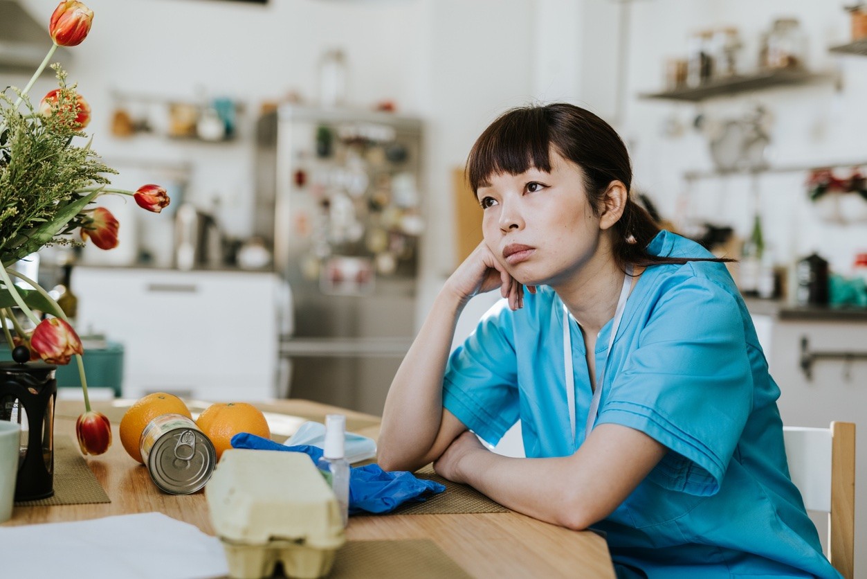 Healthcare worker in scrubs sitting at a table looking tired and thoughtful at home.