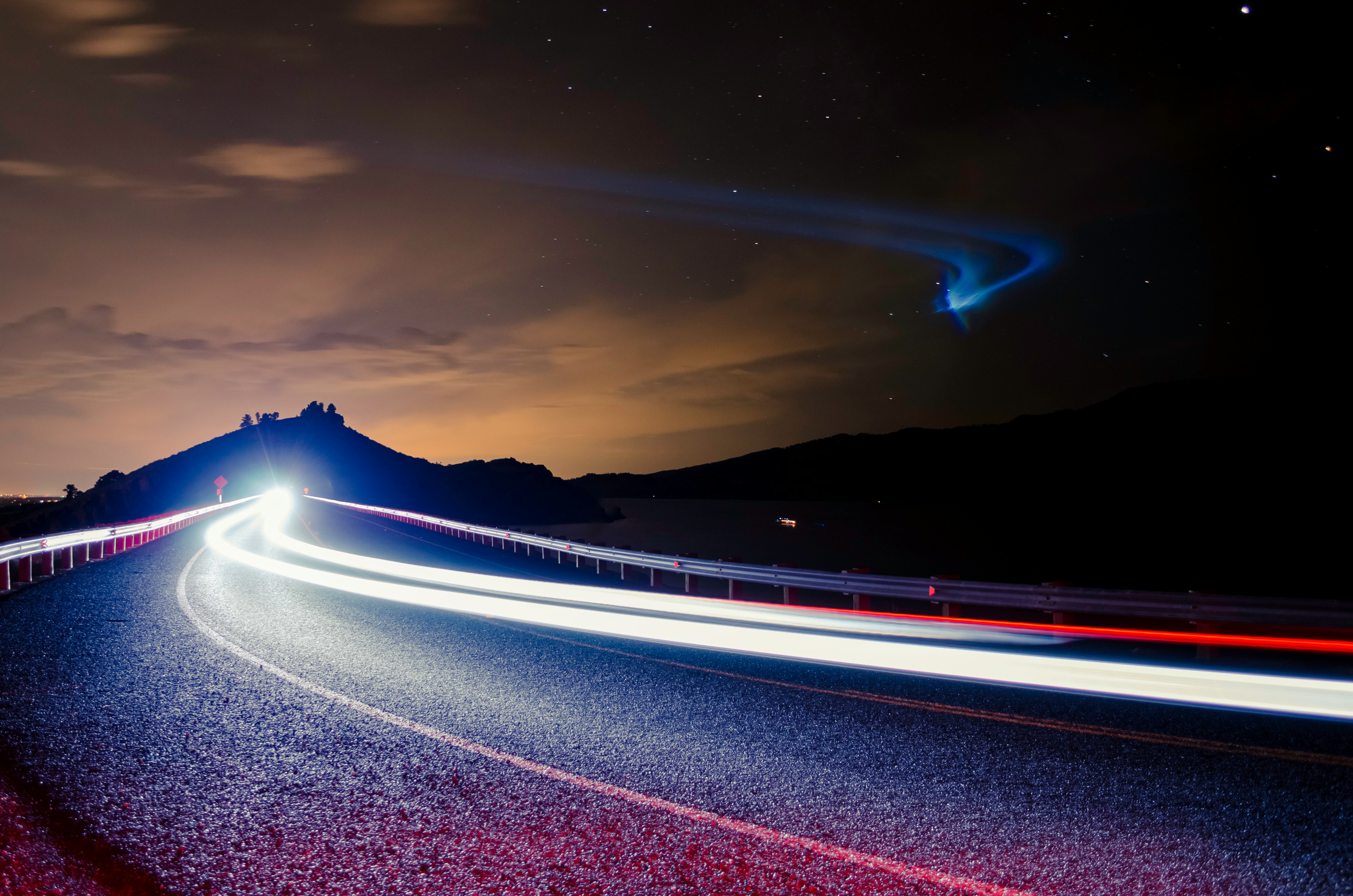 A car's headlights illuminate a winding road at night, with a vibrant sunset and distant mountains in the background.