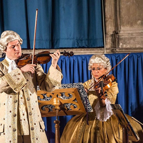 Due musicisti in costumi storici suonano il violino sul palco, con tende blu e leggii sullo sfondo.
