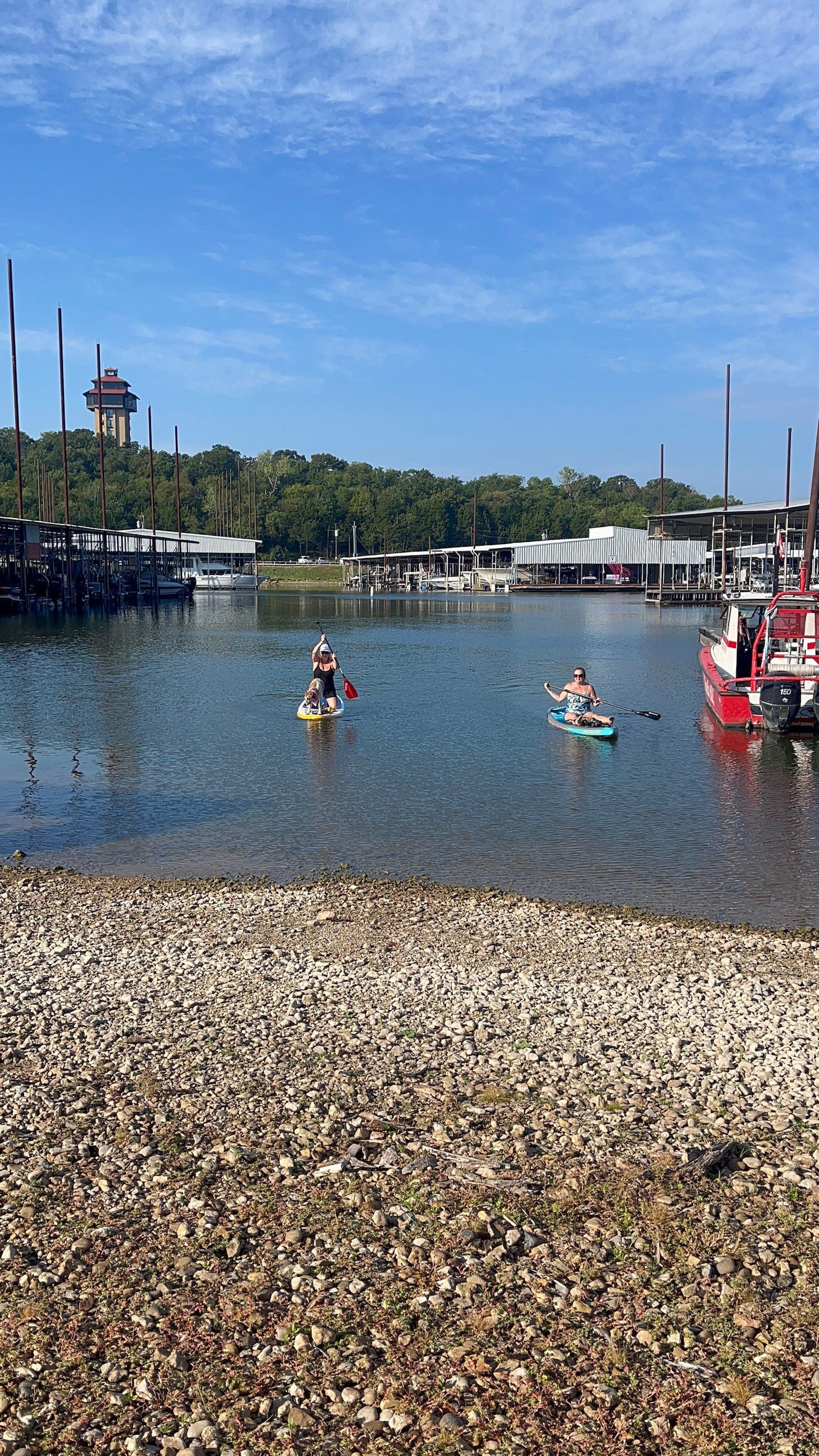 A scenic view of a clear, calm lake with two people paddleboarding near the shore, bordered by a pebble-strewn beach, with a background featuring docks, boats, and a watchtower under a vibrant blue sky.
